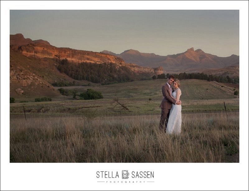 A bride and groom embracing in a golden field at sunset, dramatic red sandstone mountains stretching behind them during a farm wedding in the Cape Town region.