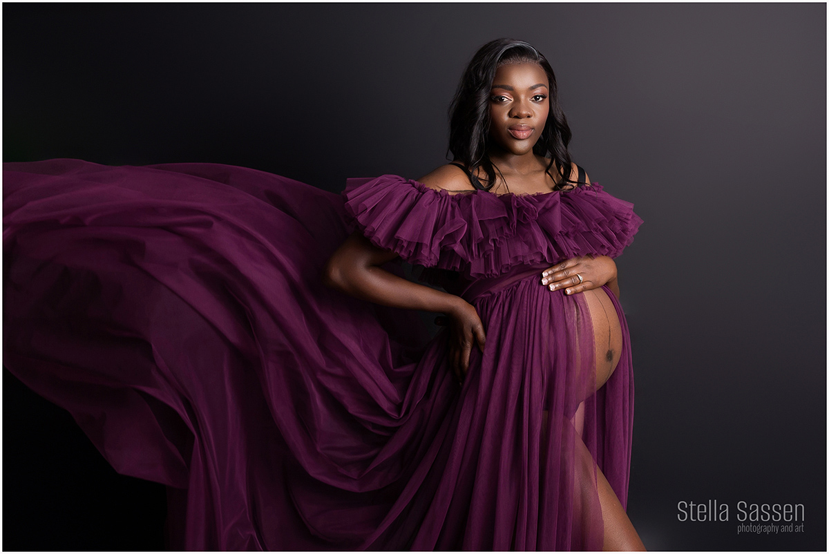 Elegant maternity portrait of a pregnant woman in a flowing deep plum gown, posing against a dark studio backdrop. The dress billows dramatically to the side, highlighting her baby bump. She gazes confidently at the camera, one hand resting gently on her belly