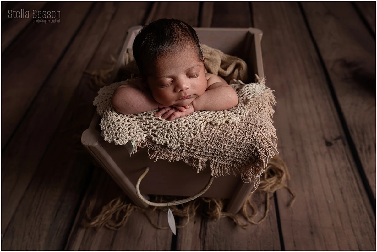 A newborn baby is posed asleep in a small wooden crate on a wooden floor backdrop. The baby is resting on their folded arms, with their head supported and facing the camera. Soft, neutral blankets and textured fabrics fill the crate, creating a warm, cosy setup.