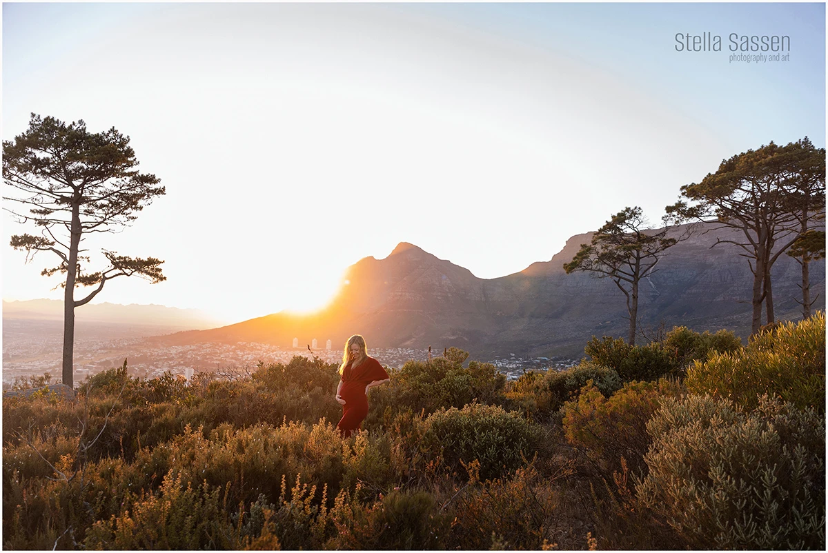 A young woman stands in greenery with Table Mountain in the background, posing for a maternity shoot